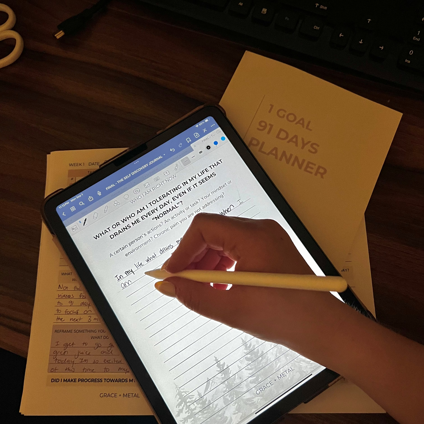 Person using a personal growth journal with a pen, on a desk with a planner and keyboard.