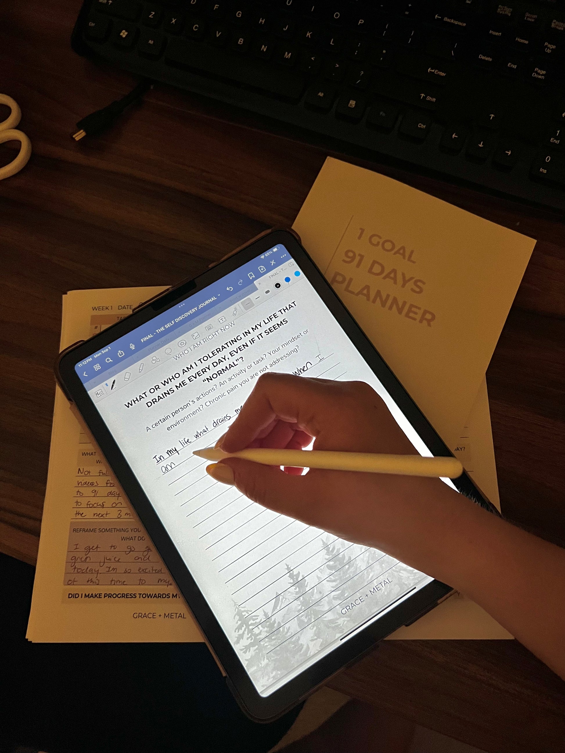 Person using a personal growth journal with a pen, on a desk with a planner and keyboard.